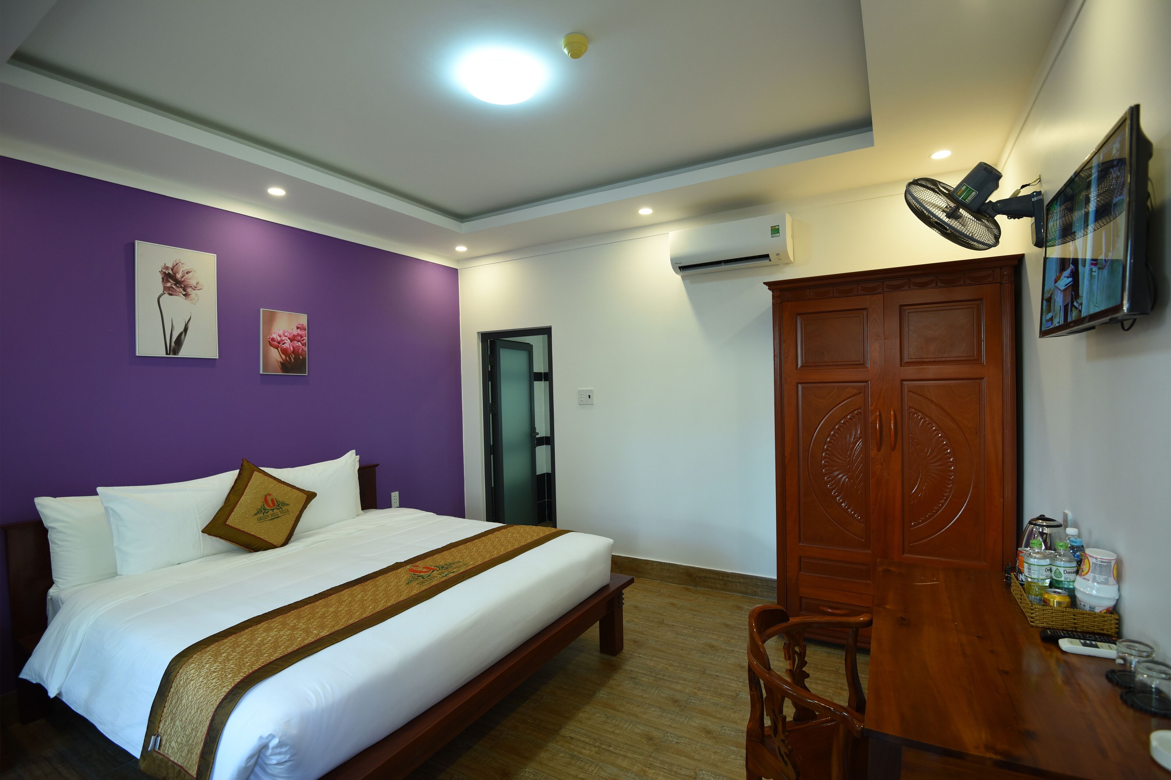 A hotel room with a bed, a wooden desk, a chair, a fan, and a door.