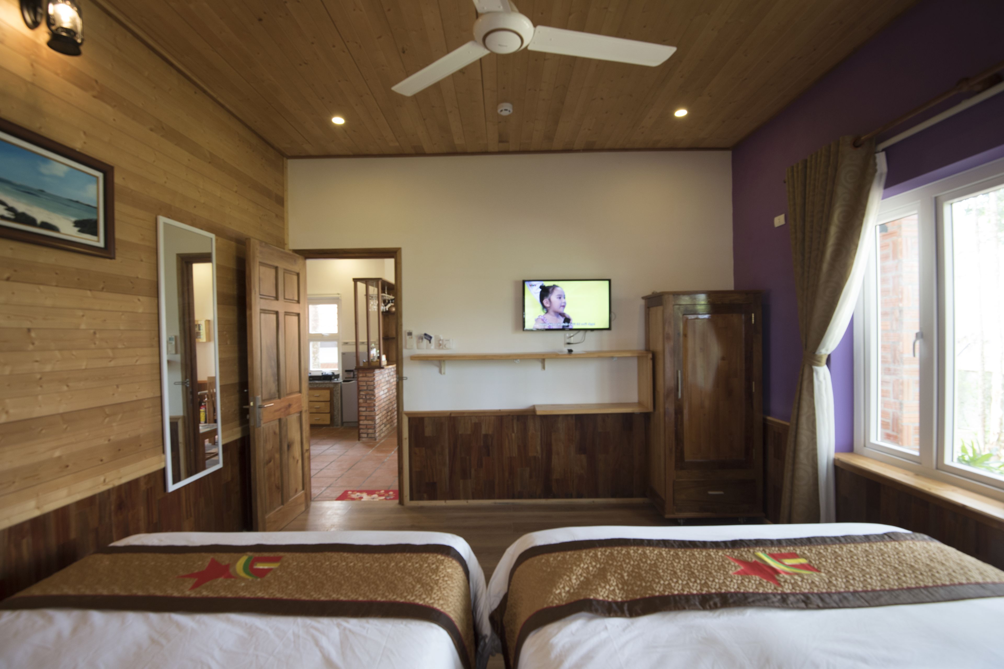 Ein Hotelzimmer mit zwei Betten, einem Fernseher, Holzwänden, einem Deckenventilator und einem Fenster mit Vorhängen.