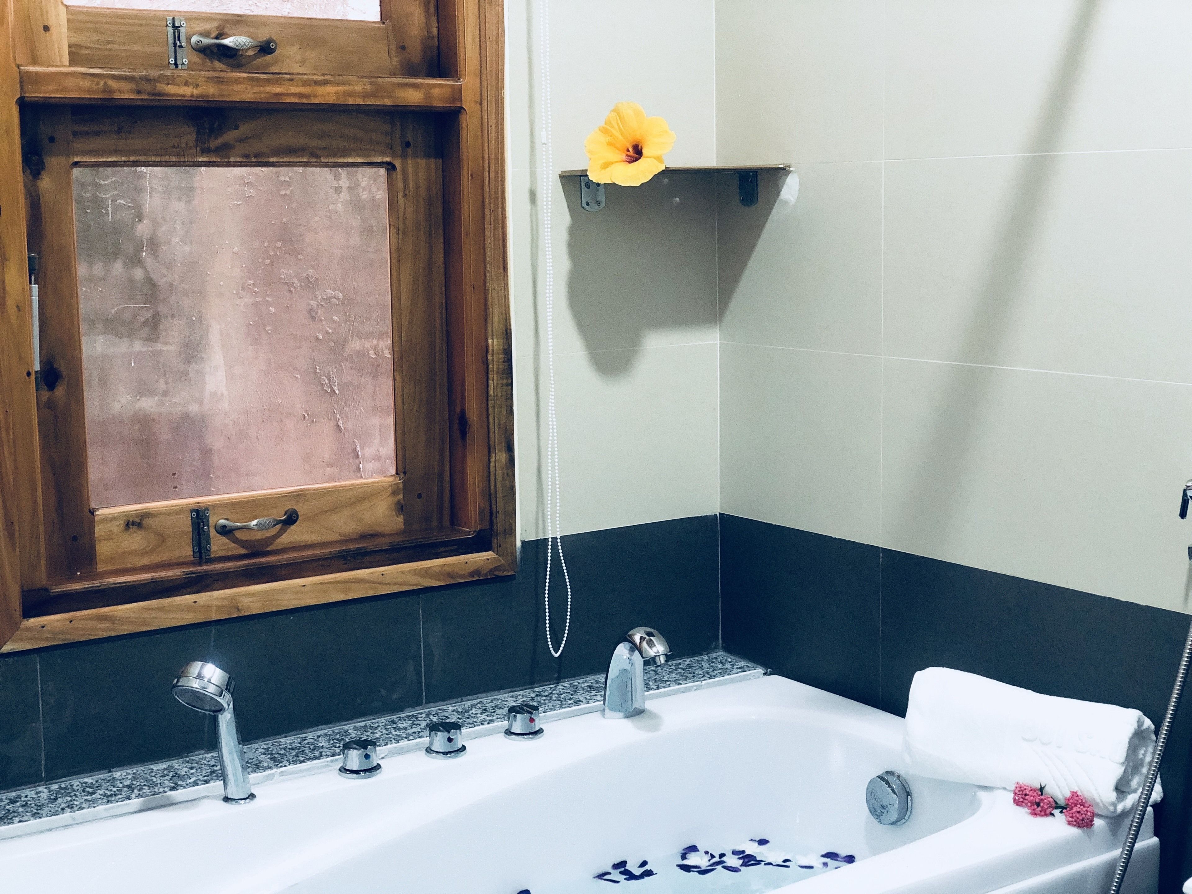 A bathroom with a wooden window, a white bathtub, a towel, and a yellow flower on a shelf.