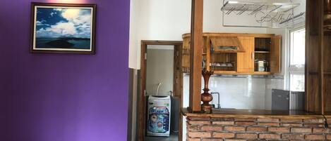 Egy lila falakkal rendelkező szoba, egy kép keretben, egy téglából épített kandalló, valamint egy konyhasarok, ahol fa szekrények és egy függesztett polc található.