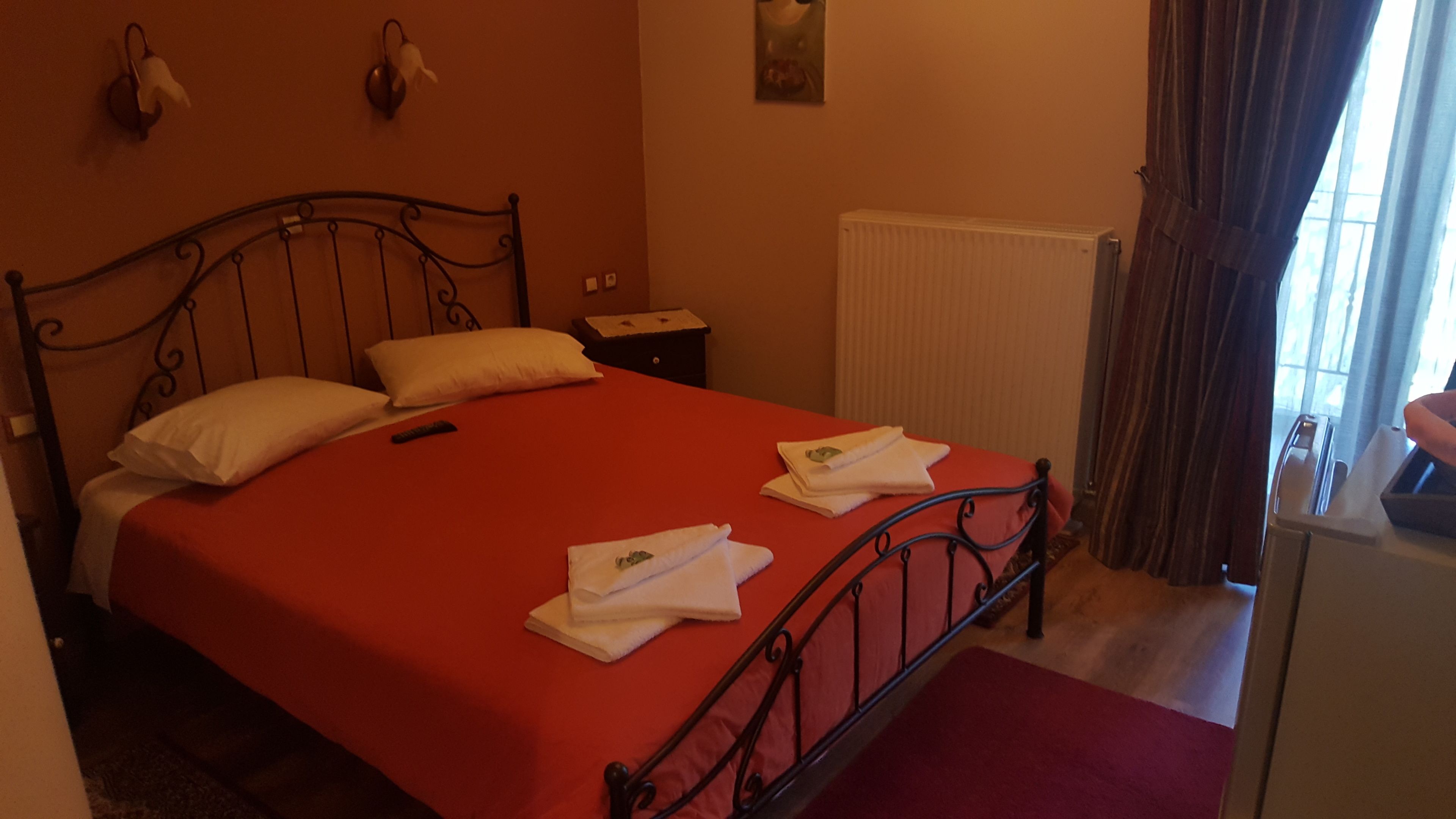 A bedroom with a red bedspread, two pillows, a nightstand, a radiator, and a window with curtains.