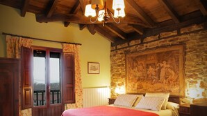 A bedroom with a stone wall, a bed with a red blanket, wooden ceiling beams, and a chandelier. - El Cajigal De Quintana (Espinosa de los Monteros)
