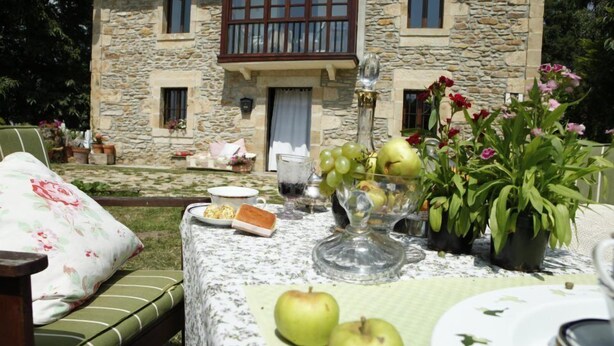 A stone building with a balcony, a table set with food and drinks, and a garden in the background. - El Cajigal De Quintana (Espinosa de los Monteros)