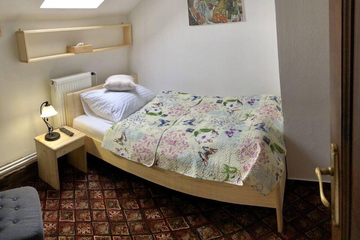 Ein kleines Schlafzimmer mit Oberlicht, einem Bett mit floralem Überwurf, einem Nachttisch mit Lampe und einem Heizkörper.