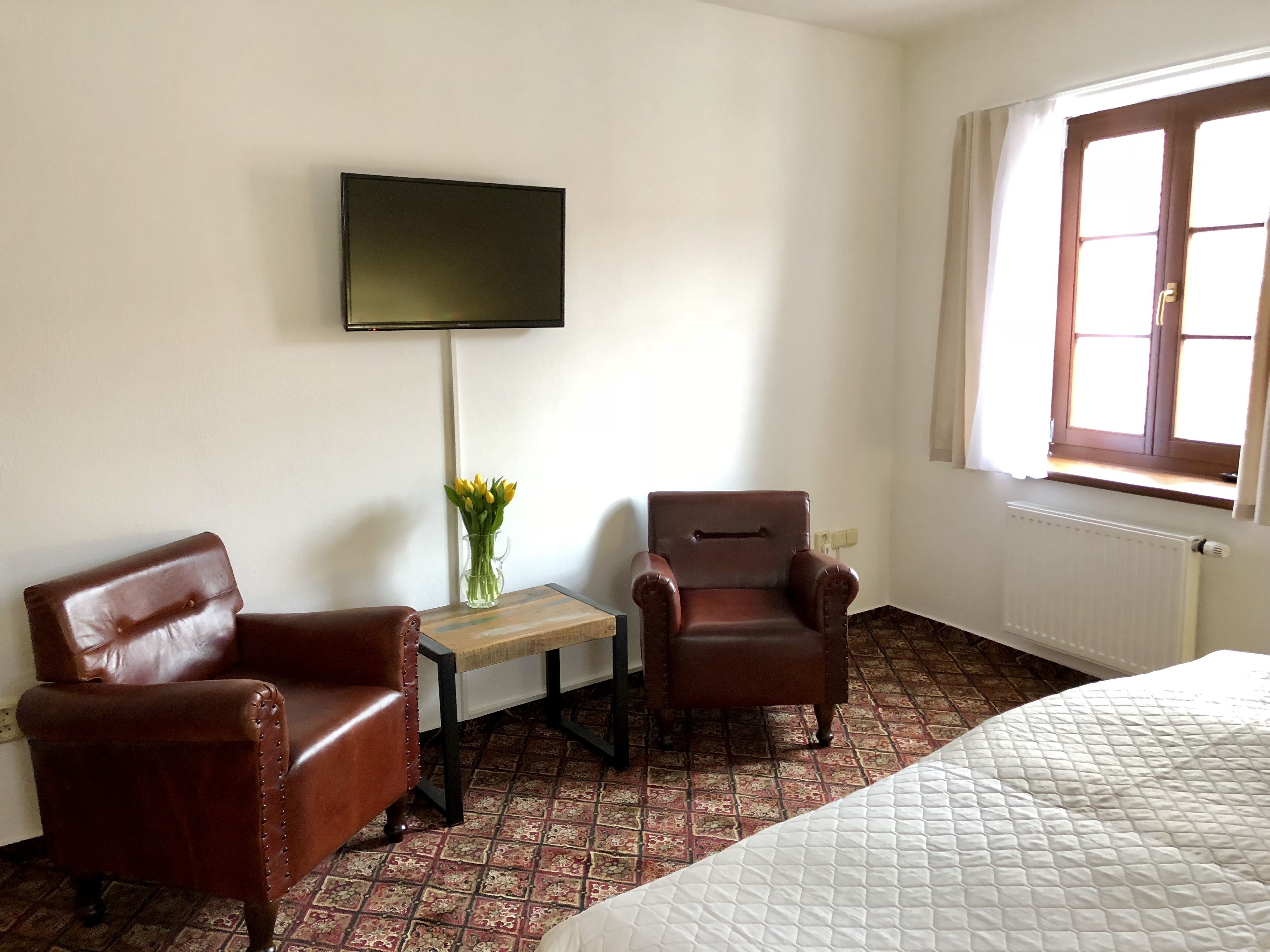 A hotel room with a bed, two leather armchairs, a small table with a vase of flowers, a flat-screen TV mounted on the wall, and a window with a view.