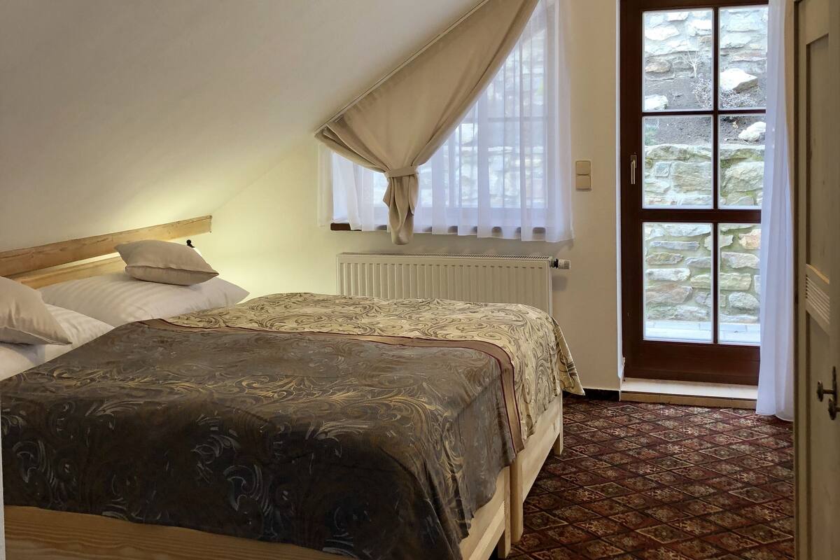 Ein Schlafzimmer mit einem großen Bett, einem Fenster mit Vorhängen, einem Heizkörper und einer zur Außenwelt führenden Tür.