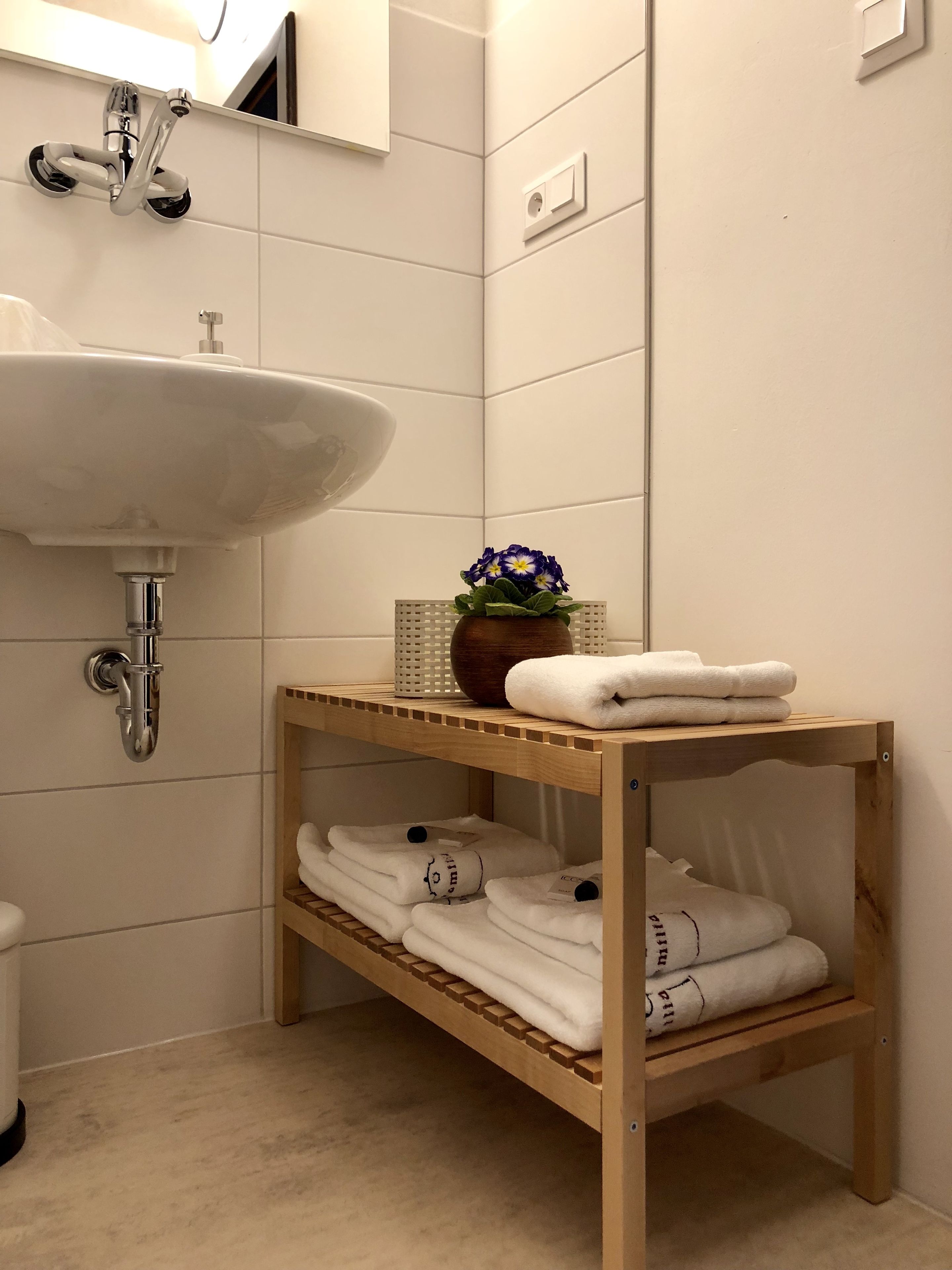 Un baño con un lavabo blanco, una estantería de madera con toallas dobladas y un jarrón con flores.