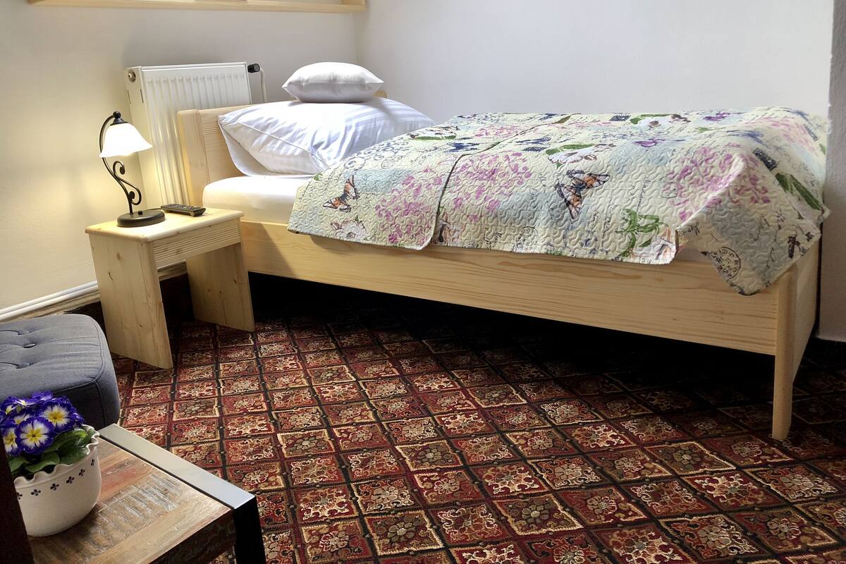 Ein kleines, ordentlich eingerichtetes Schlafzimmer mit einem Einzelbett, einem Nachttisch mit Lampe, einem Heizkörper und einem gemusterten Teppich.
