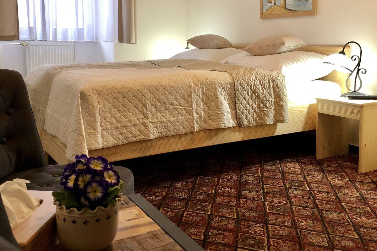 Ein Schlafzimmer mit einem Bett, einem Sessel, einem Tisch mit Blumenvase, einem Bild an der Wand und einem gemusterten Teppich.