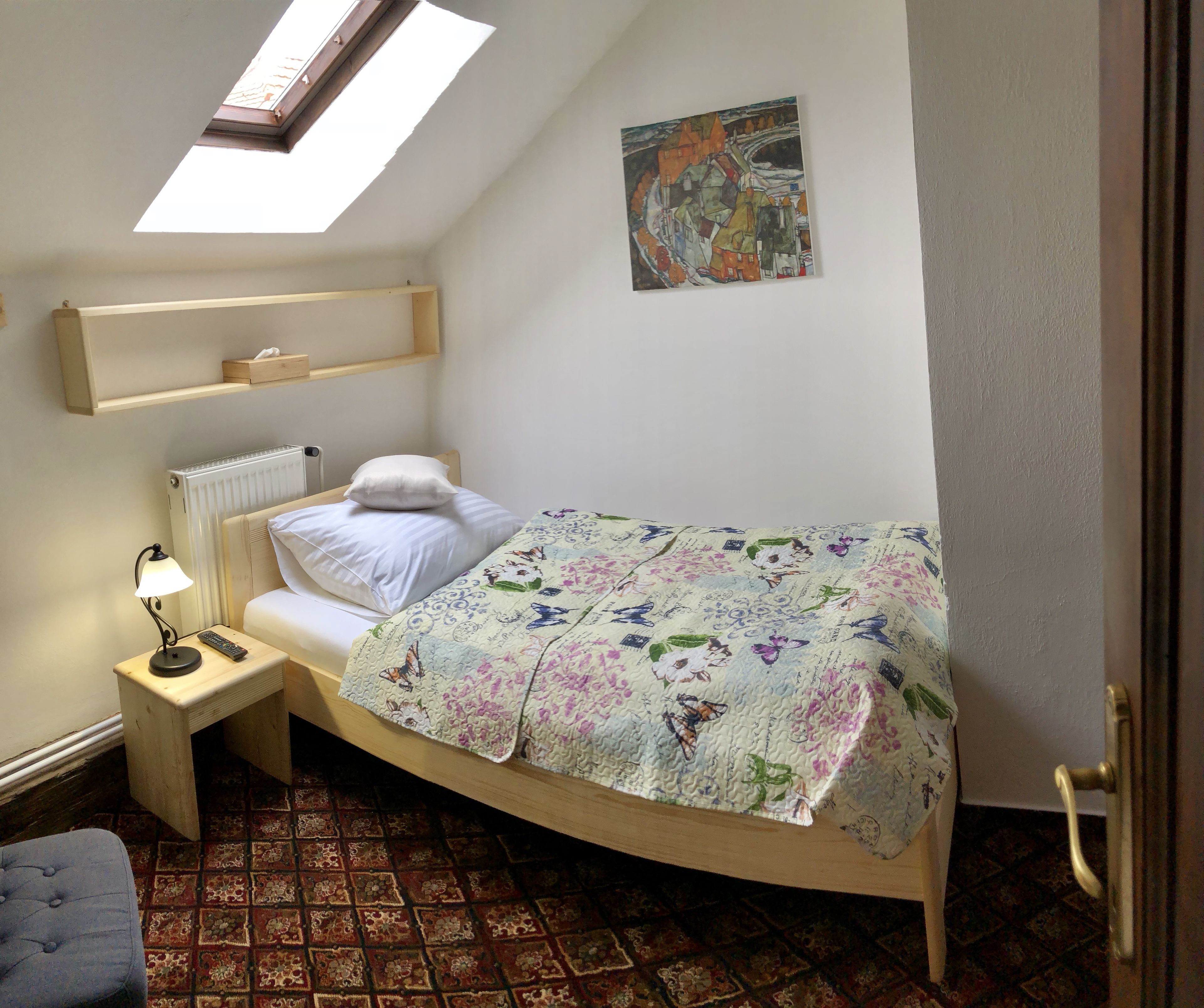 A bedroom with a skylight, a bed with a floral bedspread, a bedside table with a lamp and remote, a radiator, and a patterned rug.