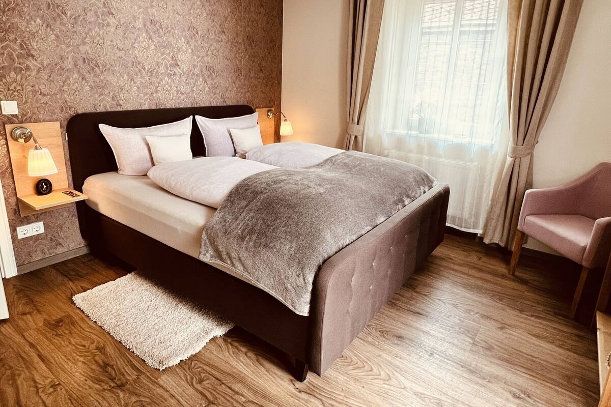 Ein ordentlich eingerichtetes Schlafzimmer mit einem großen Bett, Nachttischlampen, einem Sessel und einem Fenster mit Vorhängen.