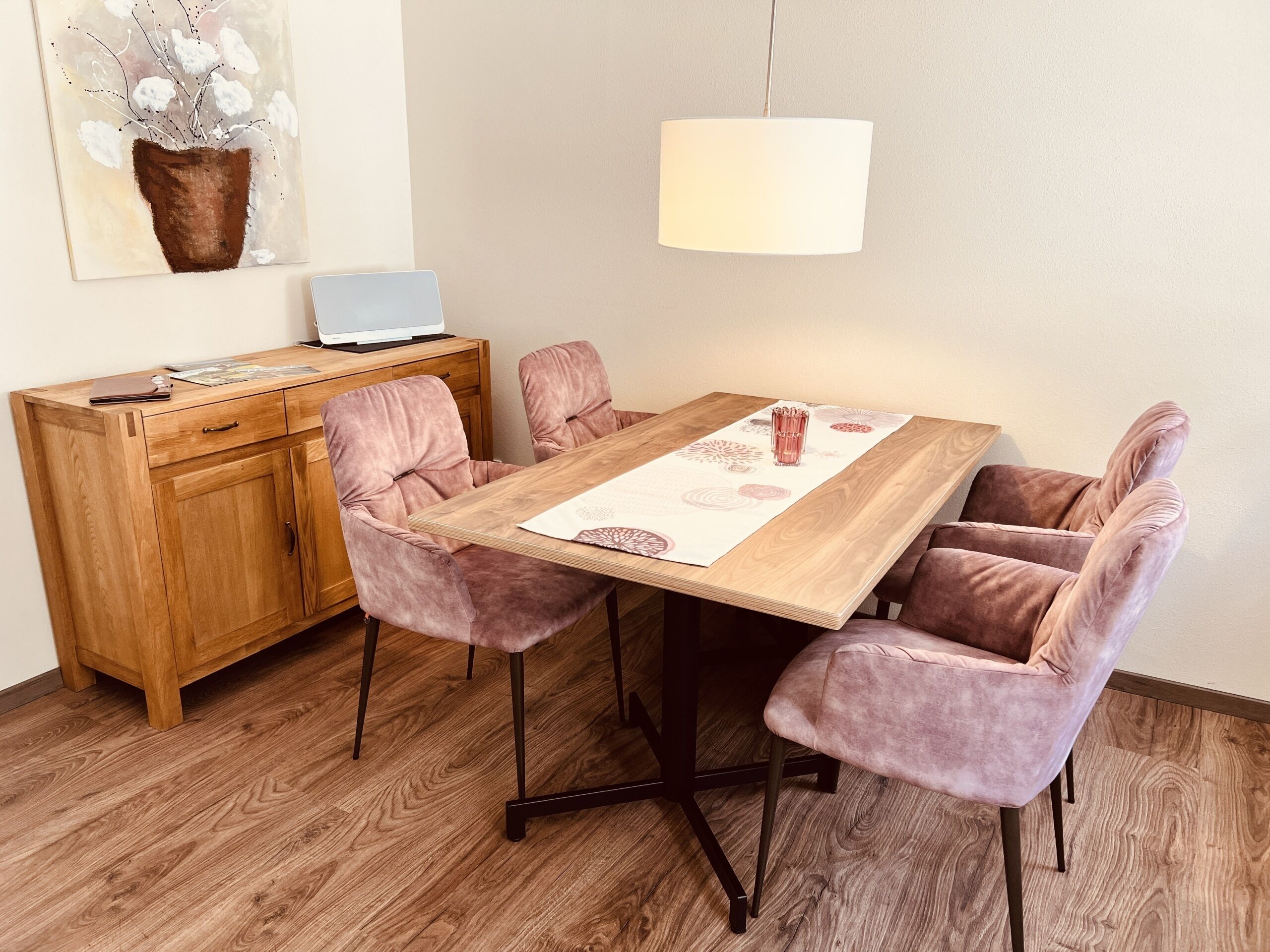 A dining area with a wooden table, four cushioned chairs, a laptop on a wooden cabinet, and a hanging light fixture.