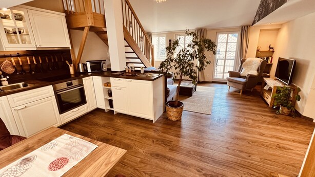 A spacious living area with a kitchen, wooden flooring, a staircase, and a large window.