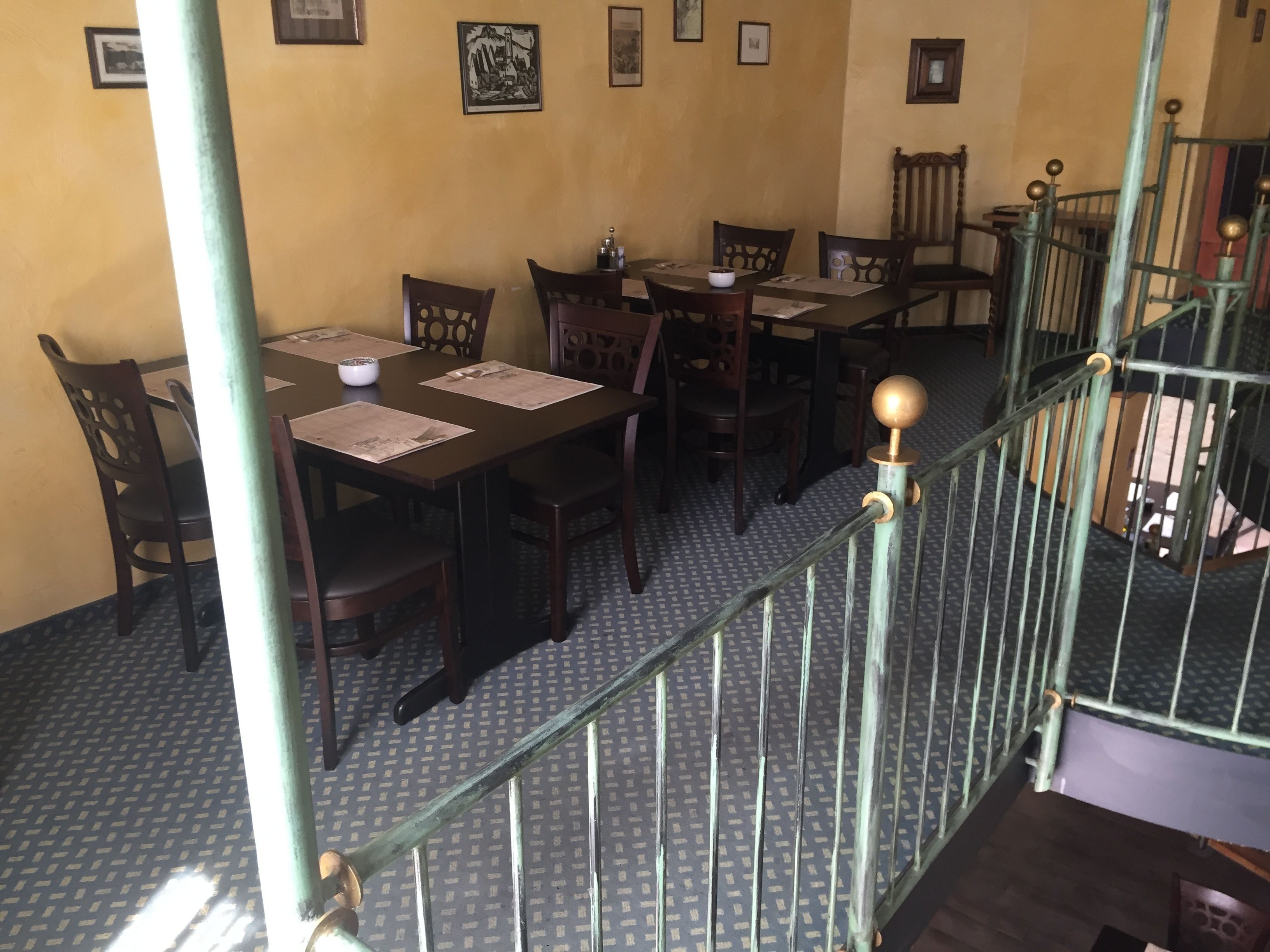 A dining area with wooden tables and chairs, a staircase with a metal railing, and patterned floor tiles.