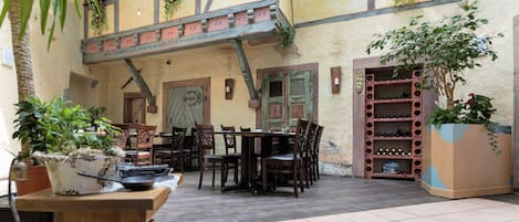 Un área de comedor al aire libre con mesas y sillas de madera, una bodega y plantas en macetas.