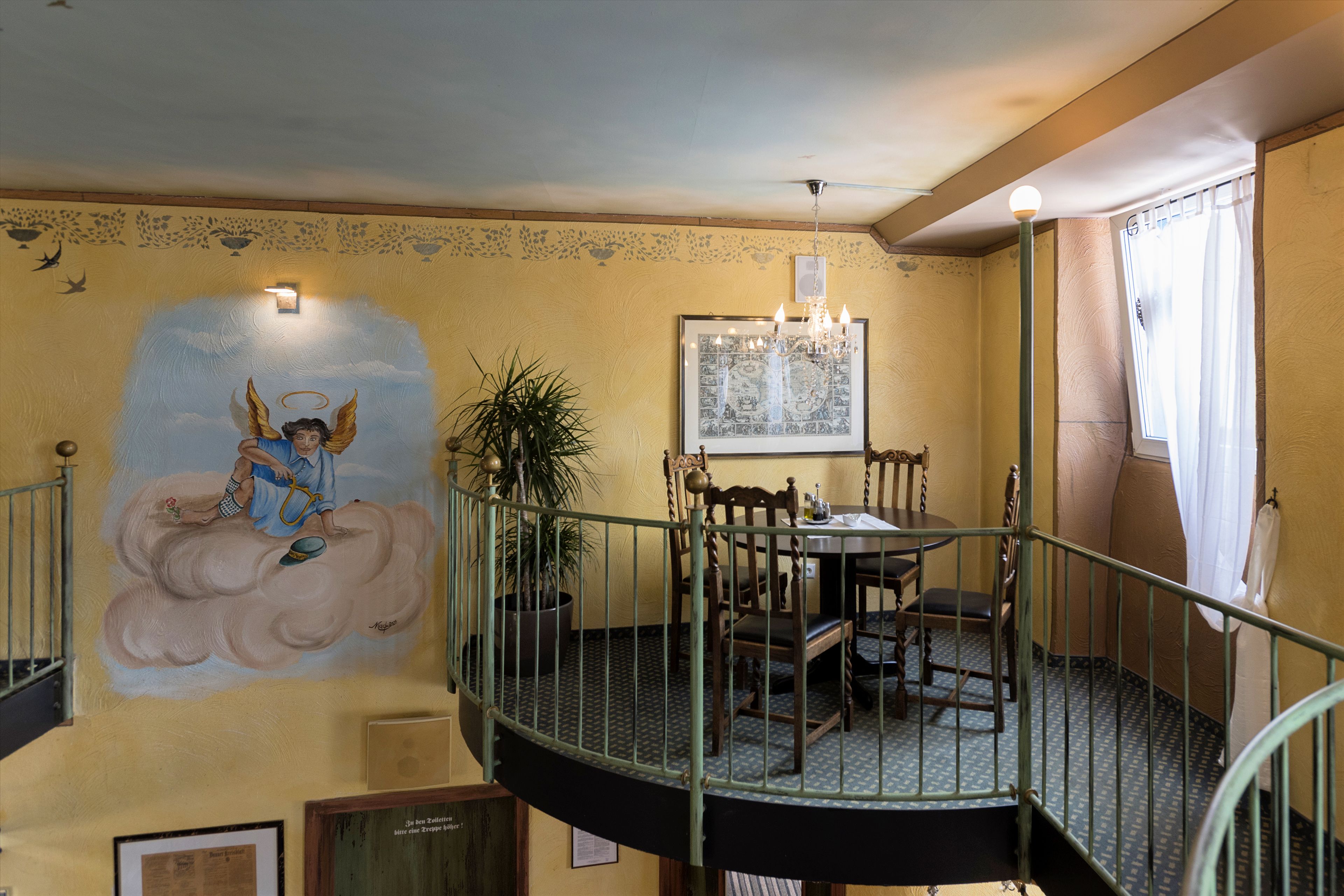 Un comedor con una mesa redonda y sillas, una araña y un mural de un ángel en la pared.