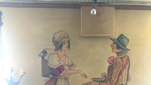 A mural of a man and woman in vintage attire, a table, and a chair. - Beim Heines (Daun)