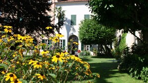A white building with green shutters, surrounded by lush greenery and yellow flowers. - Albergo Real Castello (Verduno)
