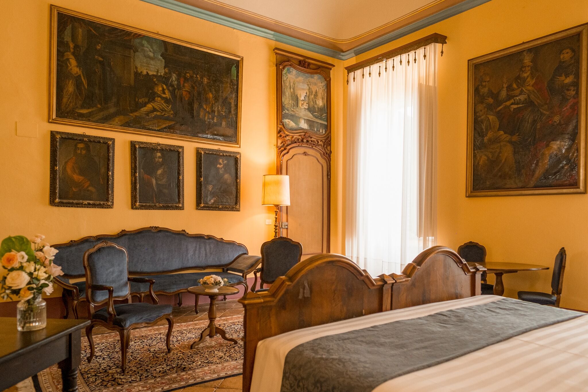 A hotel room with a large bed, a sofa, a dining table, and a chandelier. There are paintings on the walls and a window with curtains.