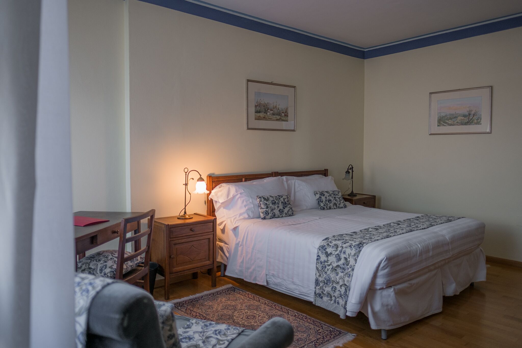 Un dormitorio con una cama doble, una mesita de noche con lámpara, una silla y un escritorio. Hay dos cuadros en la pared.