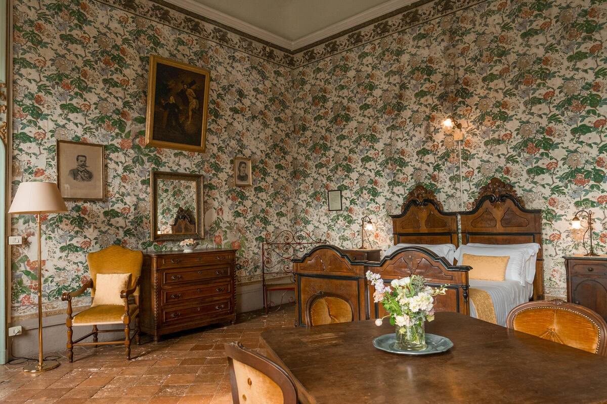 A room with floral wallpaper, a wooden dresser, a bed with a headboard, a table with a vase of flowers, and a chair.