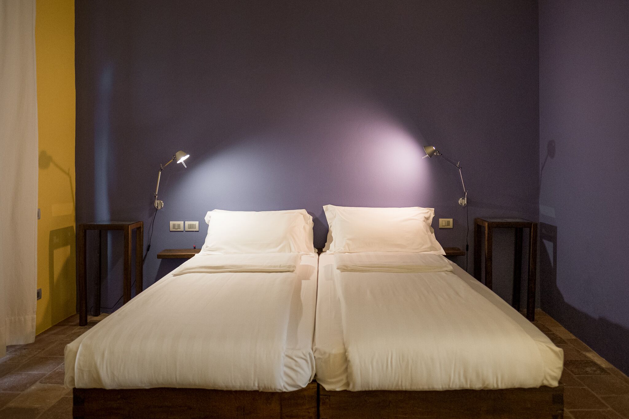 Cama doble con sábanas blancas, dos mesitas de noche y dos lámparas de pared.