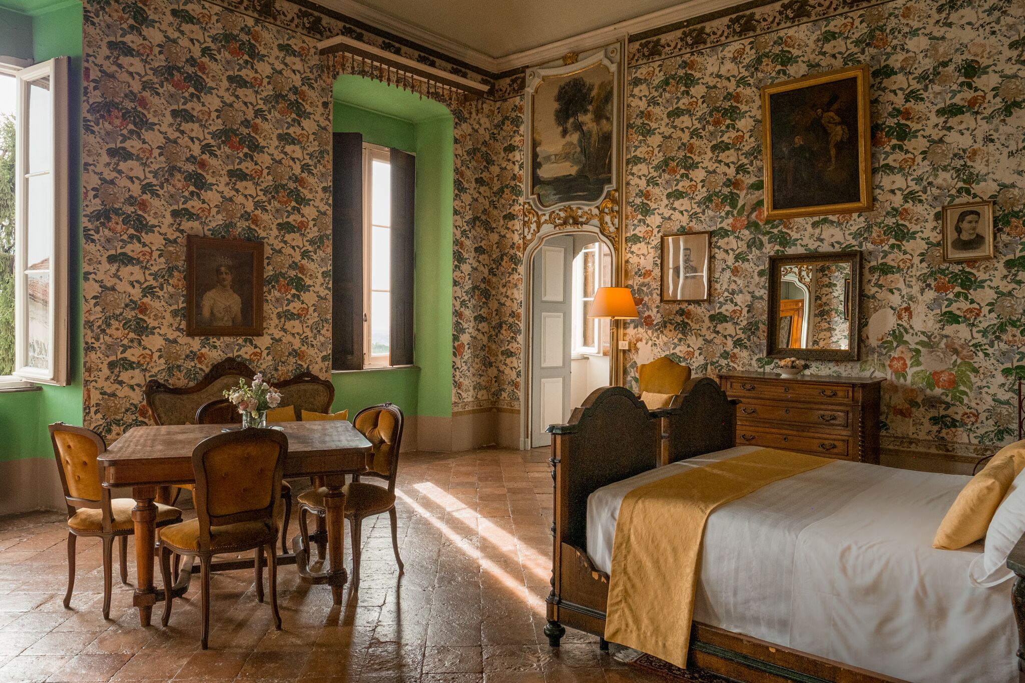 A room with floral wallpaper, a wooden bed, a dining table with chairs, and a green archway.