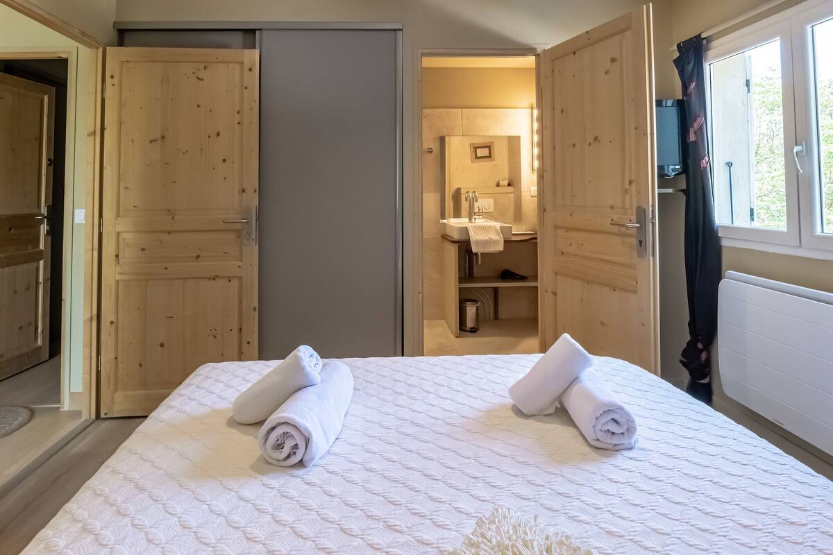 Habitació d'hotel amb un llit, dues portes de fusta, un bany amb un lavabo i un mirall, i una finestra amb cortines.