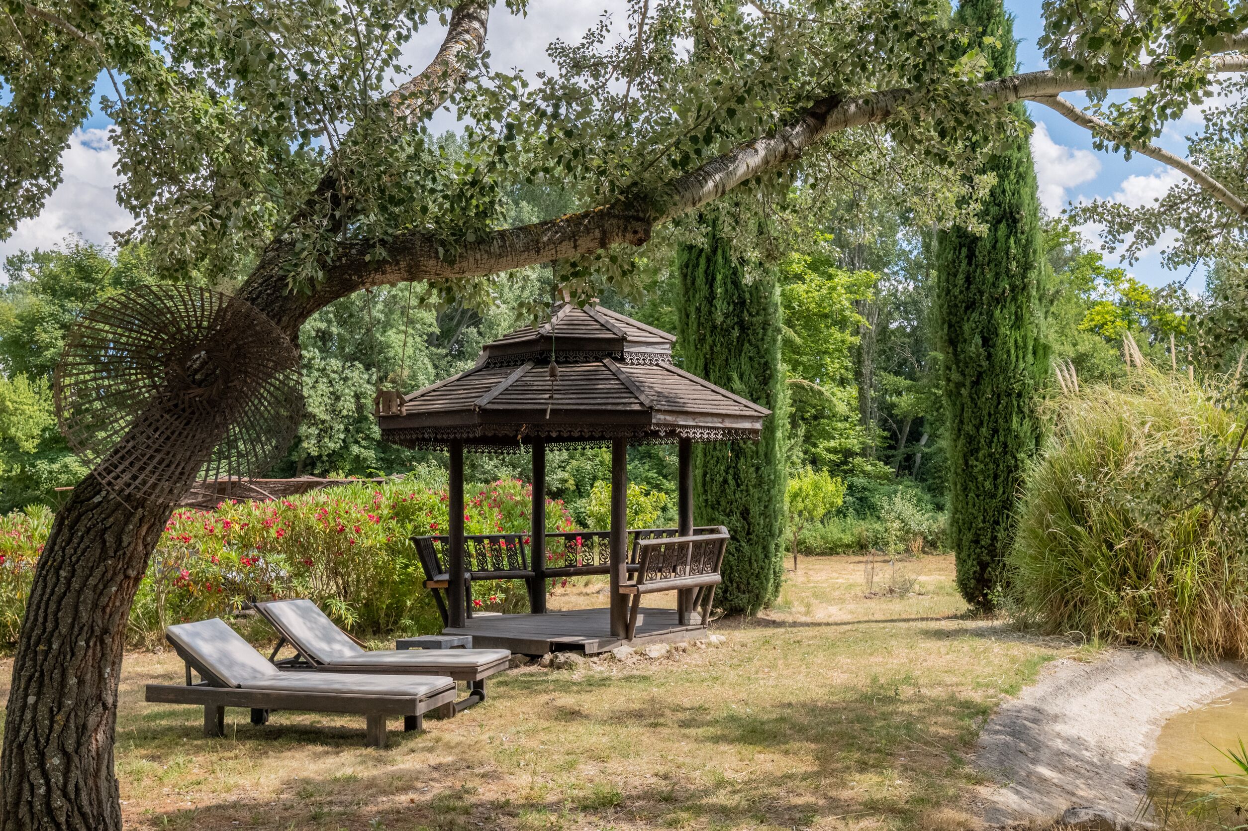 A gazebo with a roof and benches, surrounded by trees and a small pond.