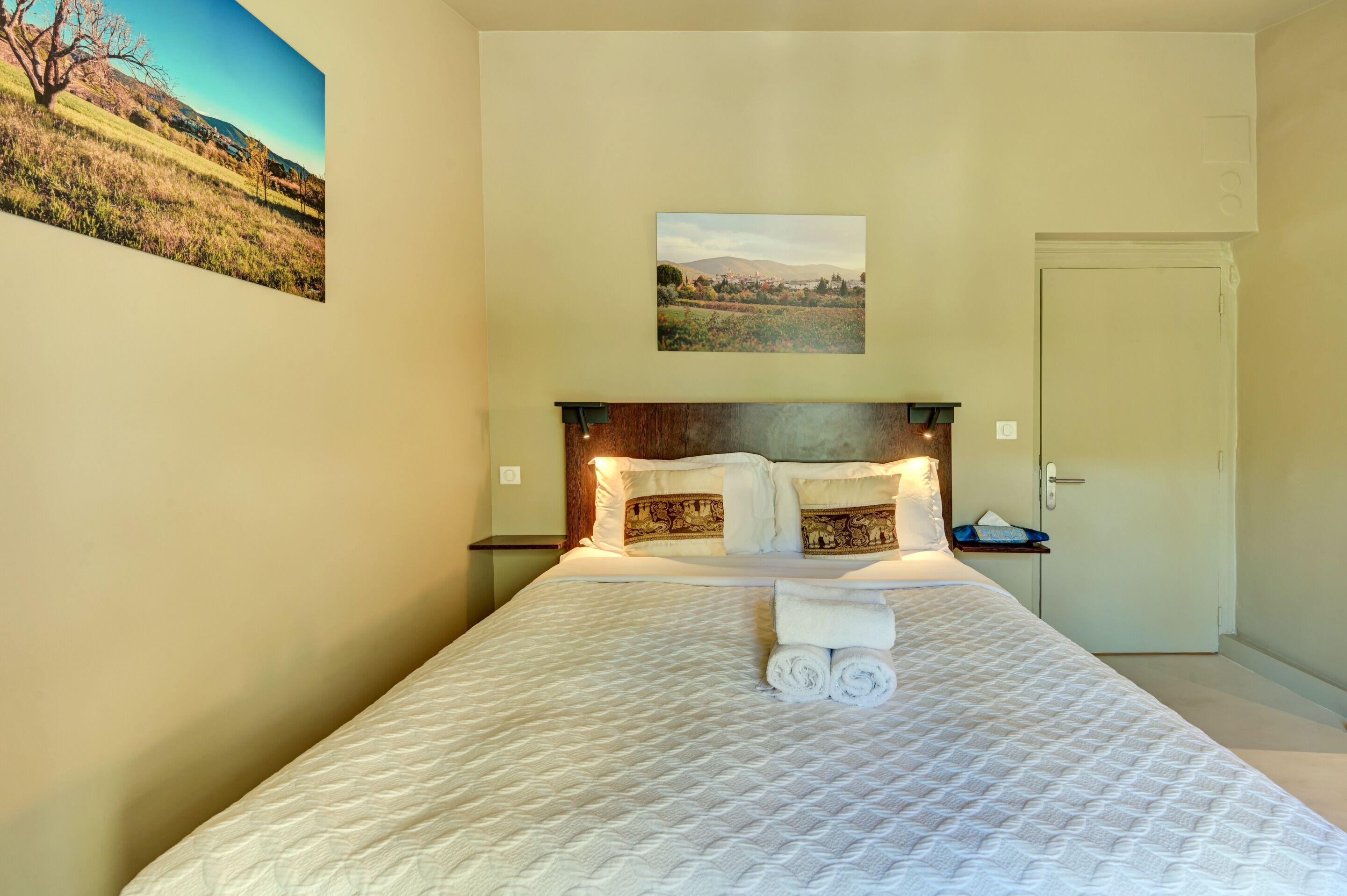 A hotel room with a bed, a headboard, a nightstand, a door, and two wall-mounted pictures.