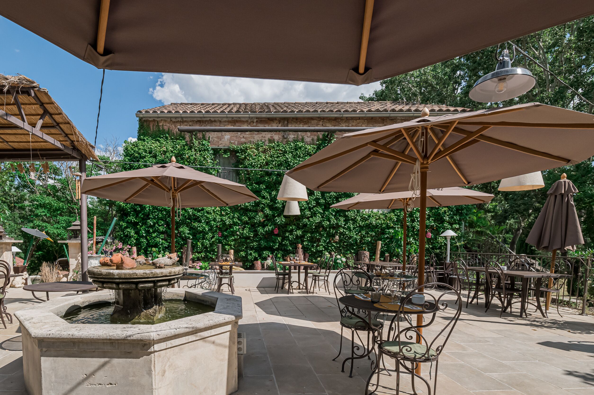 Un patio al aire libre con mesas, sillas, sombrillas, una fuente y un edificio con tejado de baldosas al fondo.