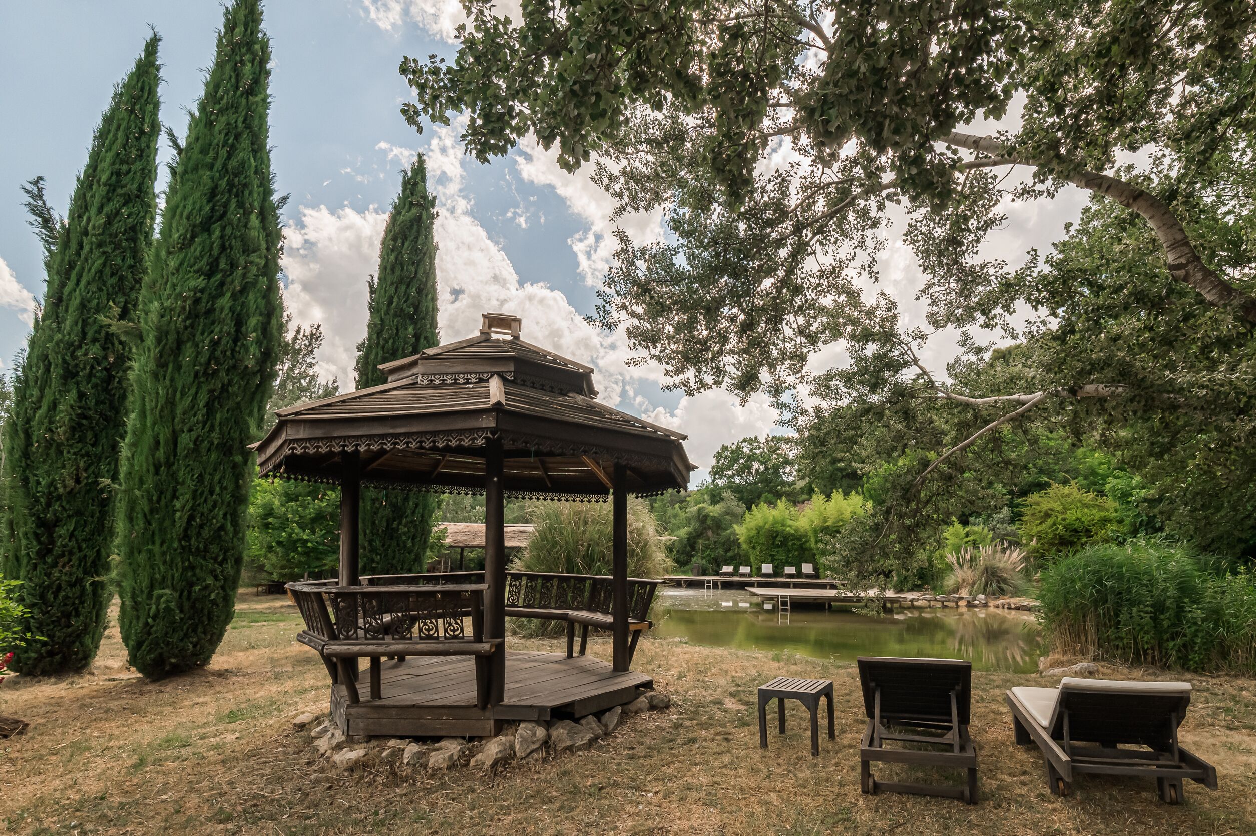 A gazebo with a wooden deck, surrounded by tall trees and a pond.