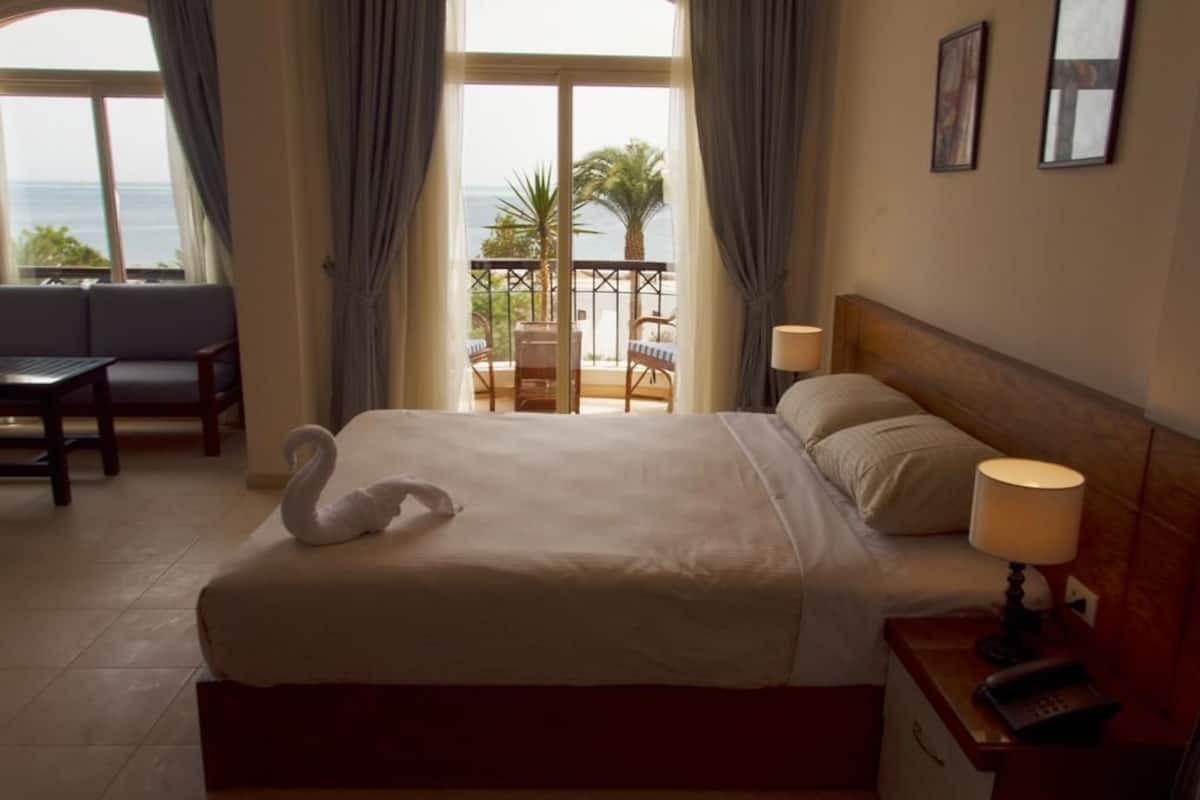 A hotel room with a large bed, a balcony with a view of the sea, a desk with a lamp, and a chair.