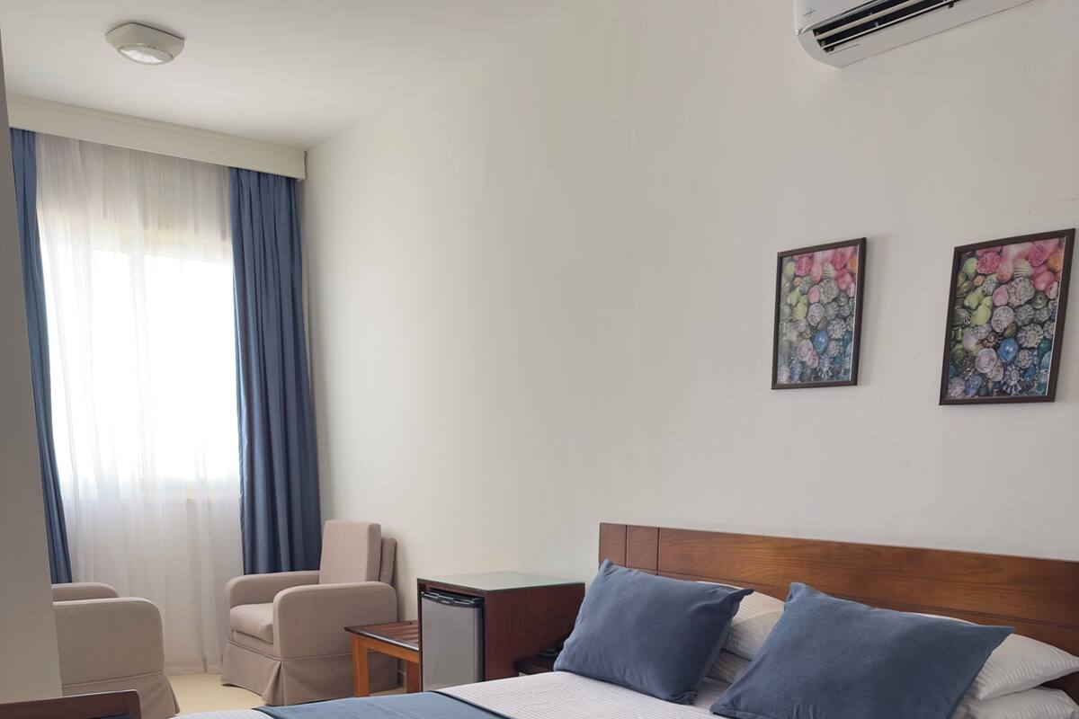 A hotel room with a bed, two chairs, a nightstand, an air conditioner, and two wall-mounted artworks.