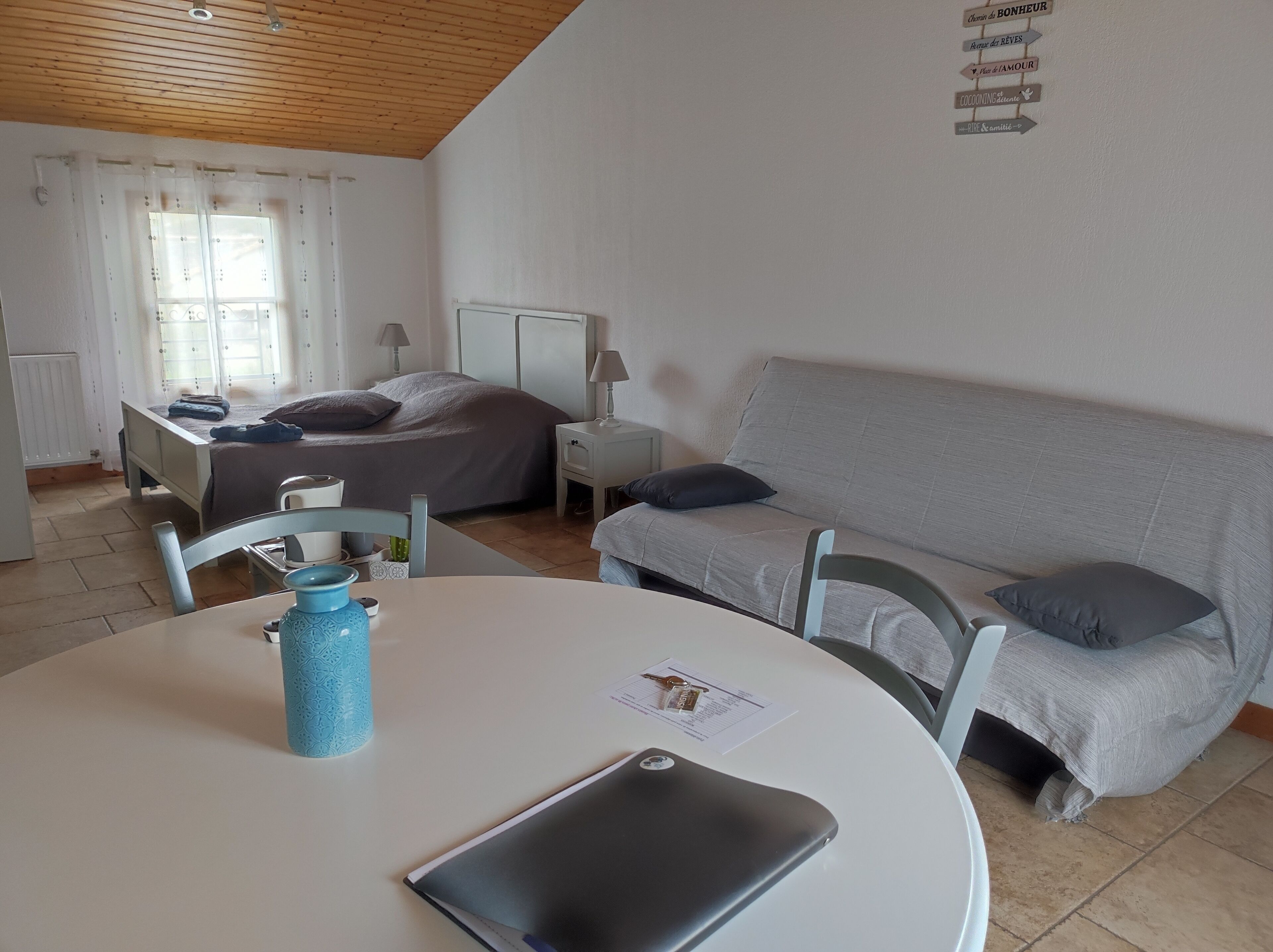 Une chambre à coucher avec un lit, un canapé gris, une table blanche avec un ordinateur portable et un vase bleu, et une fenêtre avec des rideaux.