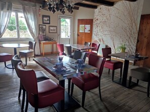 A dining area with wooden tables and chairs, a view of the outdoors through a window, and a wall with a tree branch mural. - Les Chenêts (Chenay)