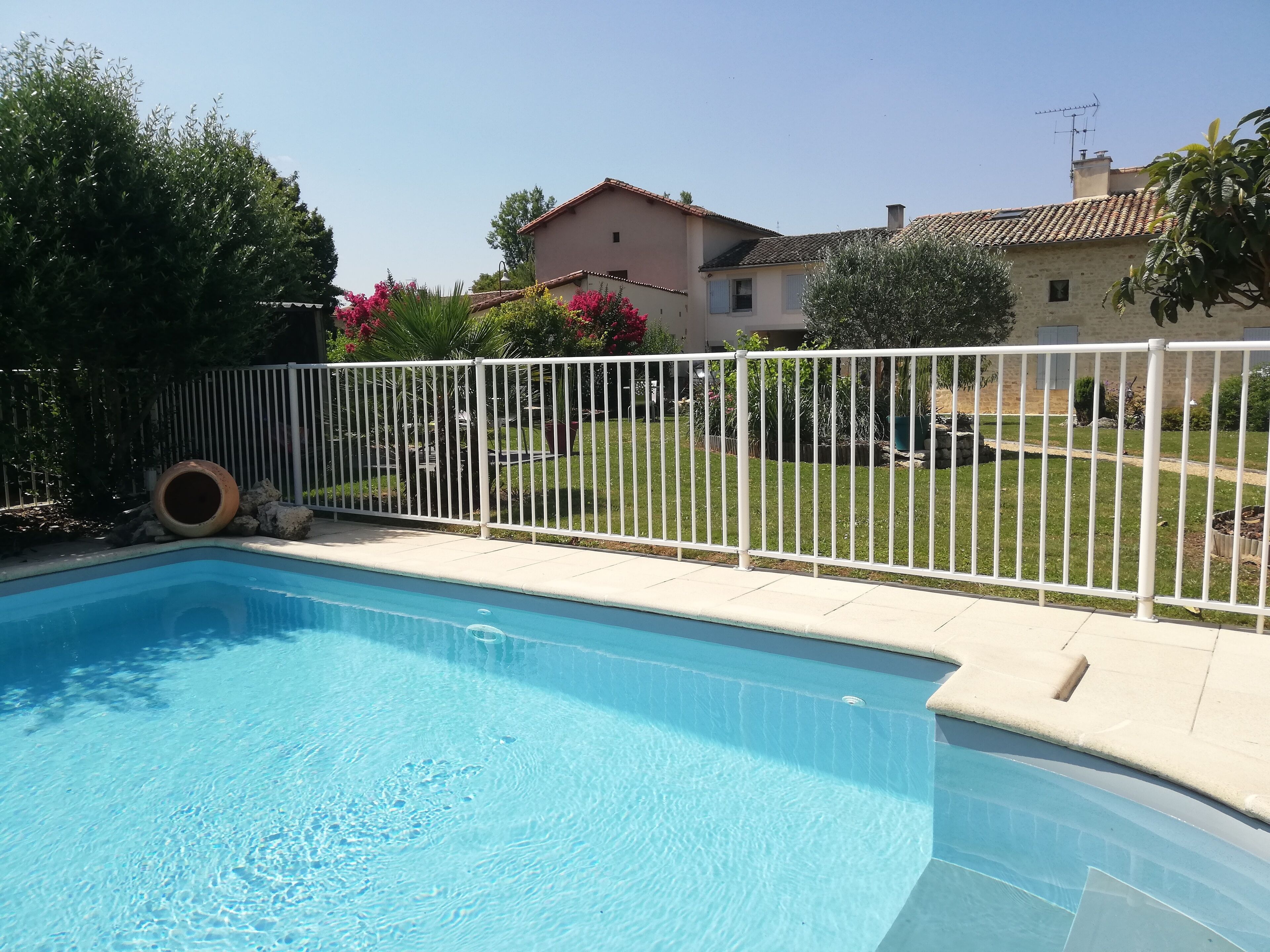 A swimming pool with a white fence, surrounded by a grassy yard and a house with a tiled roof.