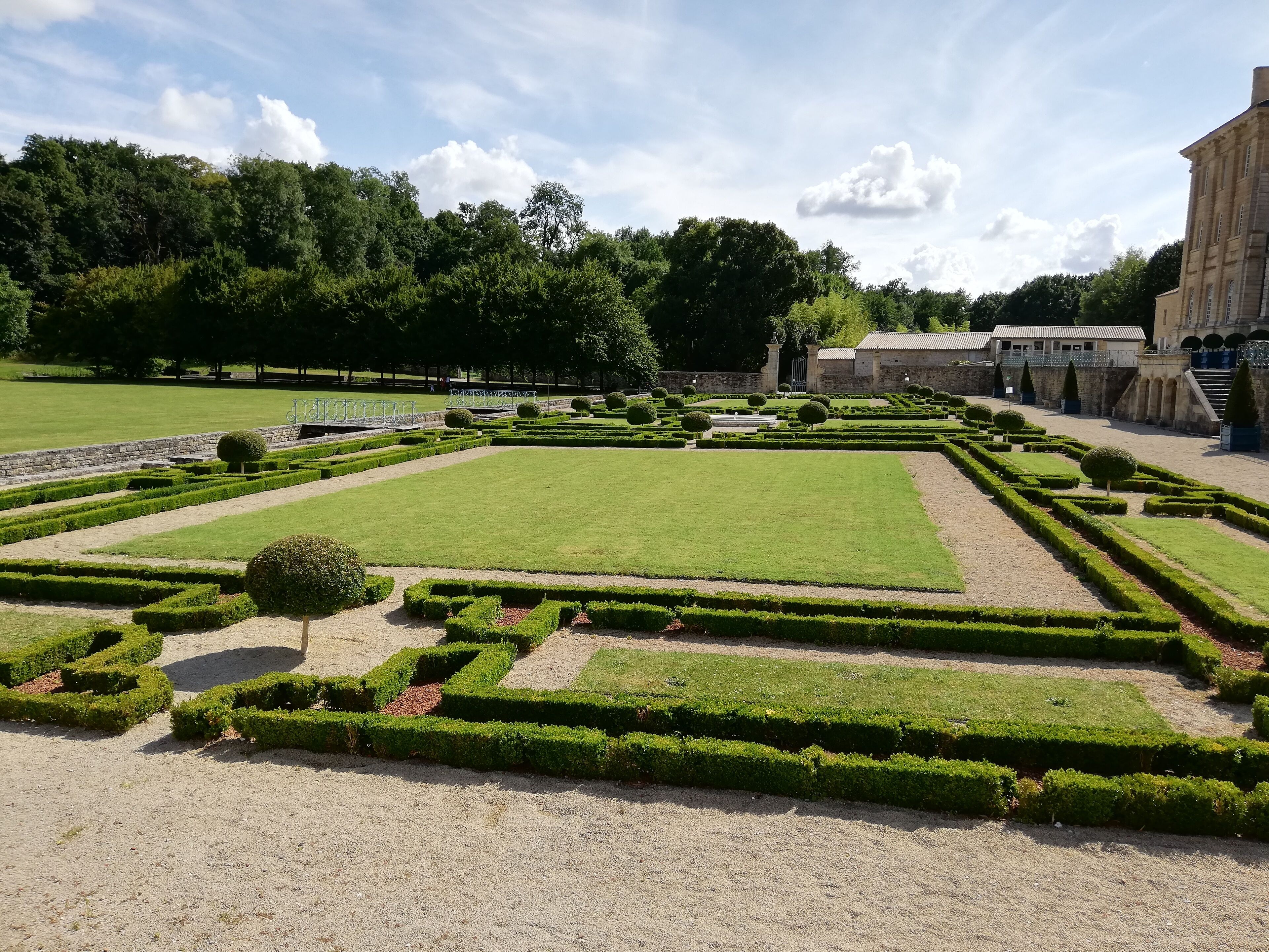 Un jardin à la française, avec des haies aux formes géométriques, une pelouse centrale et un bâtiment de style classique en arrière-plan.