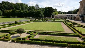 Un jardin à la française, avec des haies aux formes géométriques, une pelouse centrale et un bâtiment de style classique en arrière-plan.