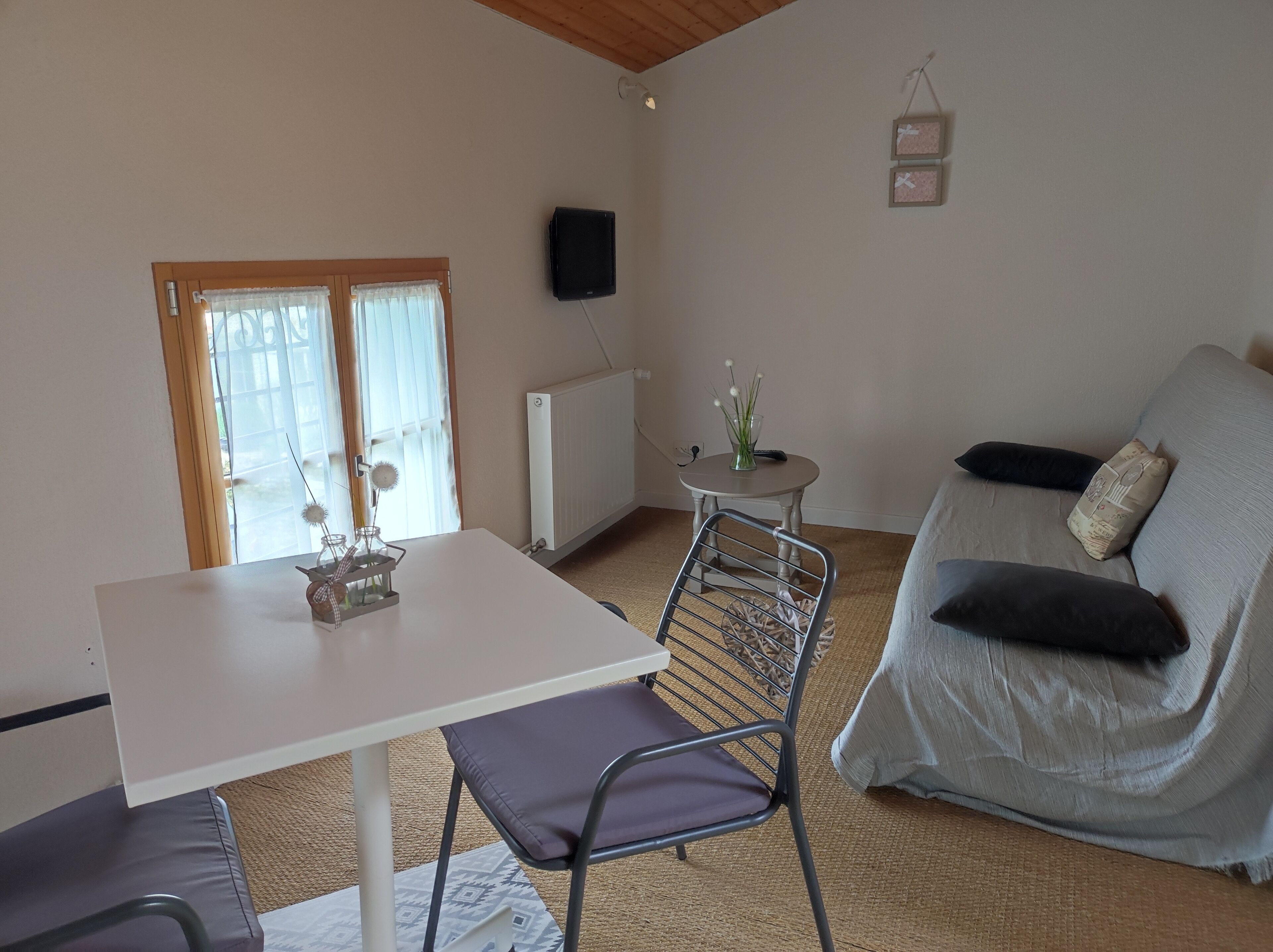 Ein gemütliches Wohnzimmer mit einem weißen Tisch, einem grauen Sofa mit Kissen, einem kleinen runden Tisch mit einer Vase und einer Heizung.