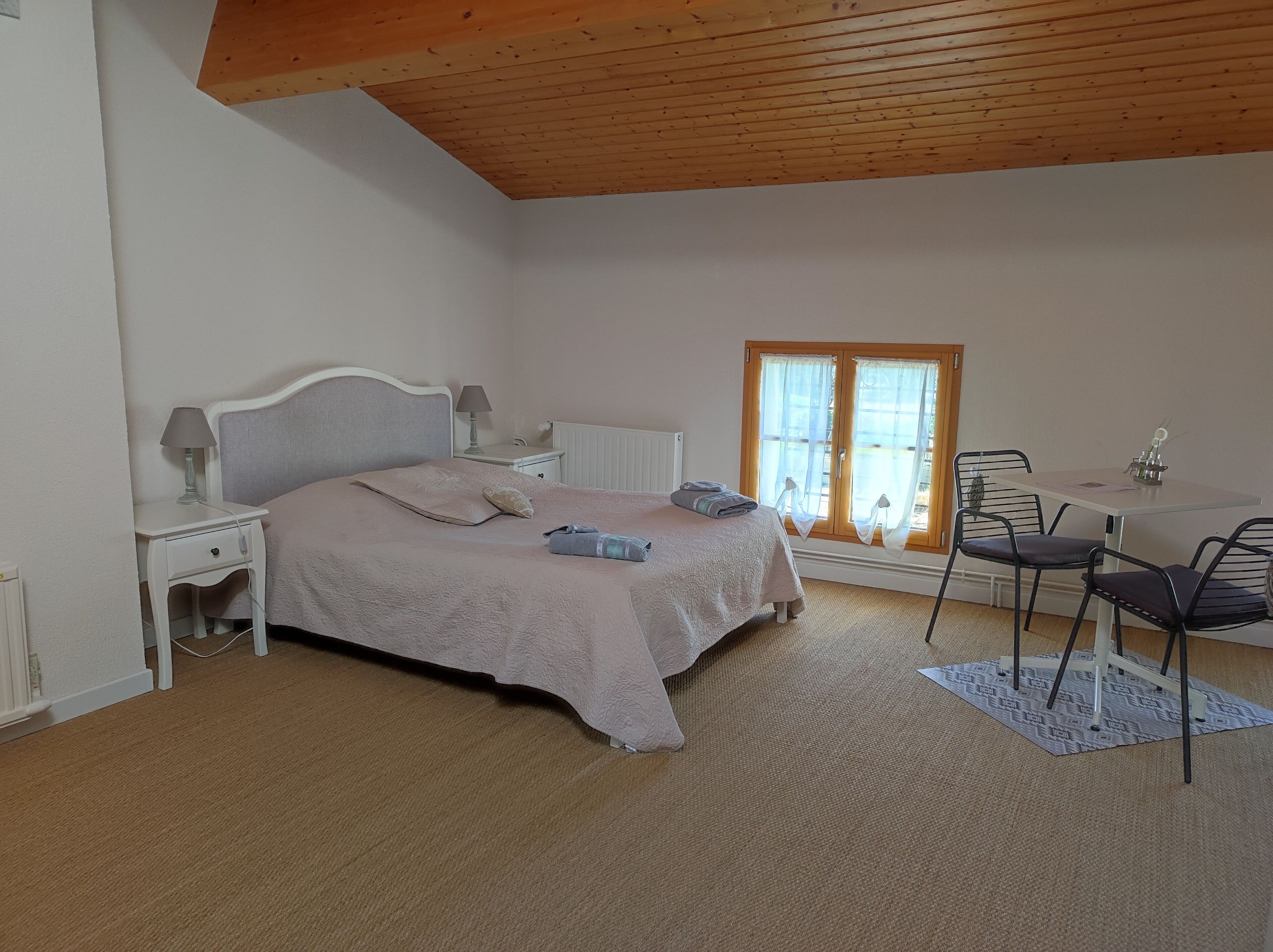 Quarto com cama, mesas de cabeceira, abajur, janela com cortinas e uma pequena mesa com cadeiras.