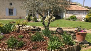 A stone building with a tiled roof, a well-maintained garden with various plants and flowers, and a small tree in the center. - Les Chenêts (Chenay)