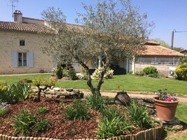 Un bâtiment en pierre avec un toit en tuiles, un jardin bien entretenu avec diverses plantes et fleurs, et un petit arbre au centre.