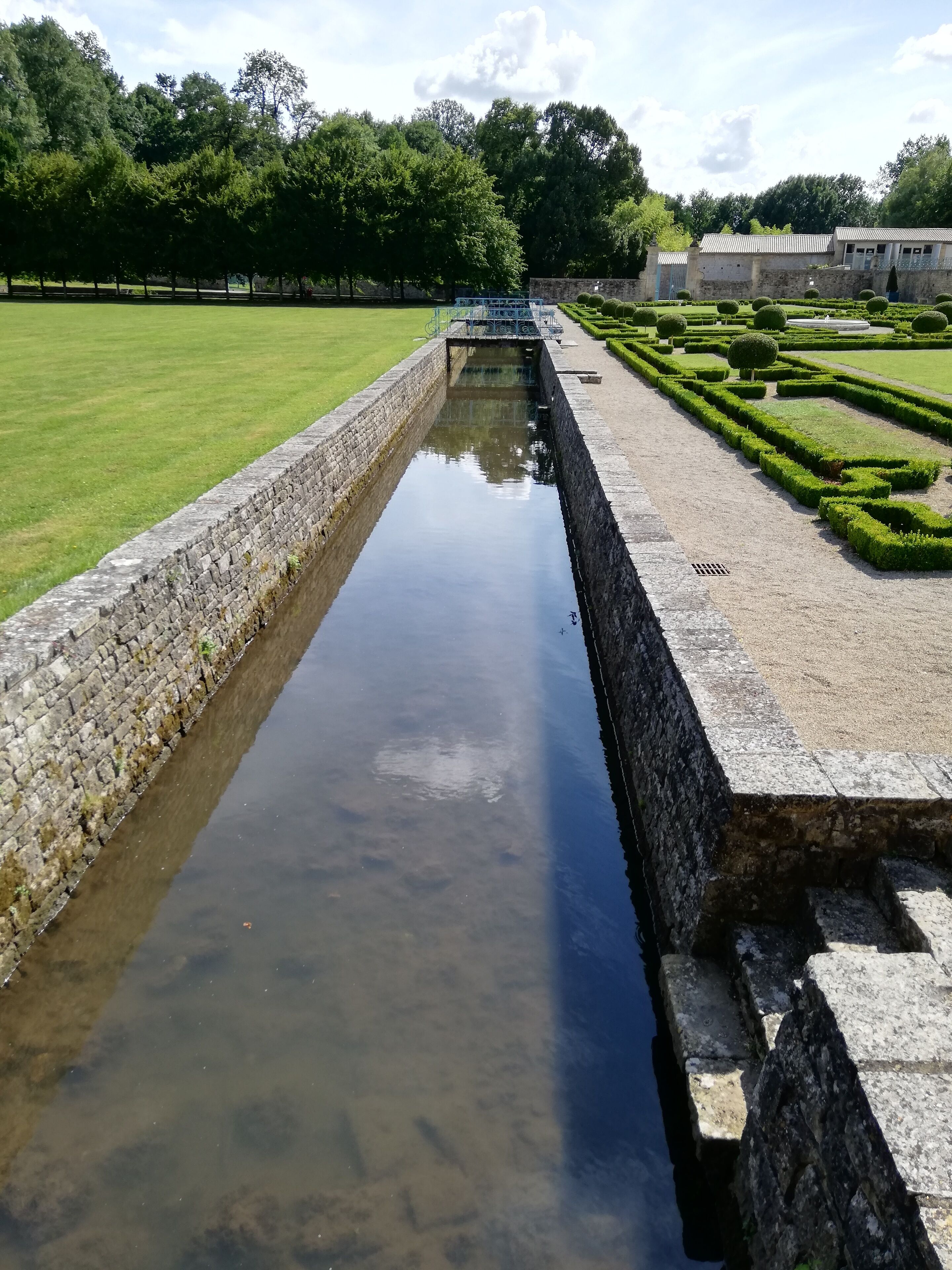 Un canal rectangular de agua con paredes de piedra, rodeado por un césped cuidado y un jardín formal con setos recortados y árboles.