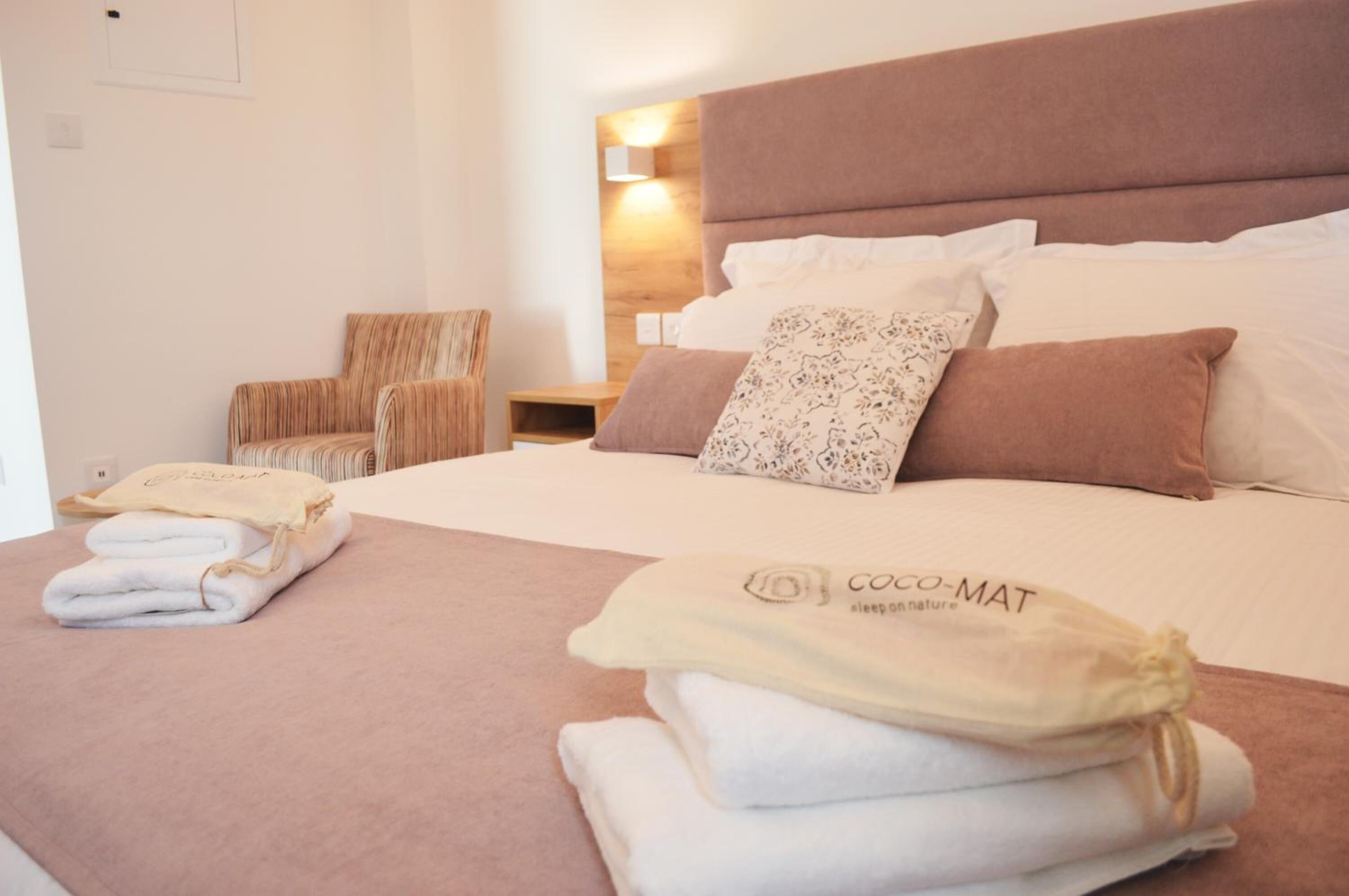 Un lit bien fait, avec du linge de lit blanc et marron, une table de chevet avec une lampe et une chaise agrémentée d’un coussin à rayures.