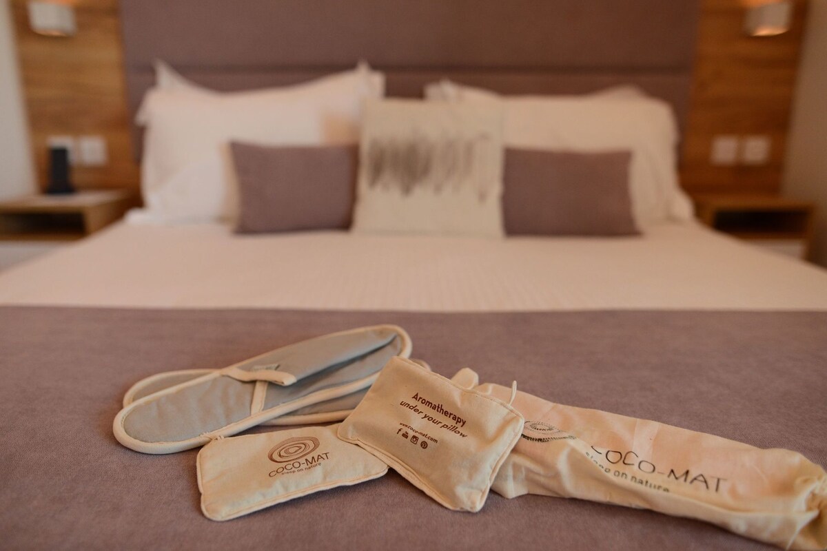 Een netjes opgemaakt bed met kussens en dekbed, een paar slippers en een opgerolde handdoek met tekst erop.