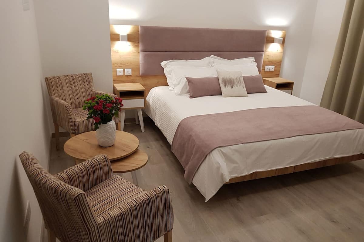 Een hotelkamer met een groot bed, twee nachtkastjes, een ronde houten tafel, twee gestreepte stoelen en een vaas met bloemen.