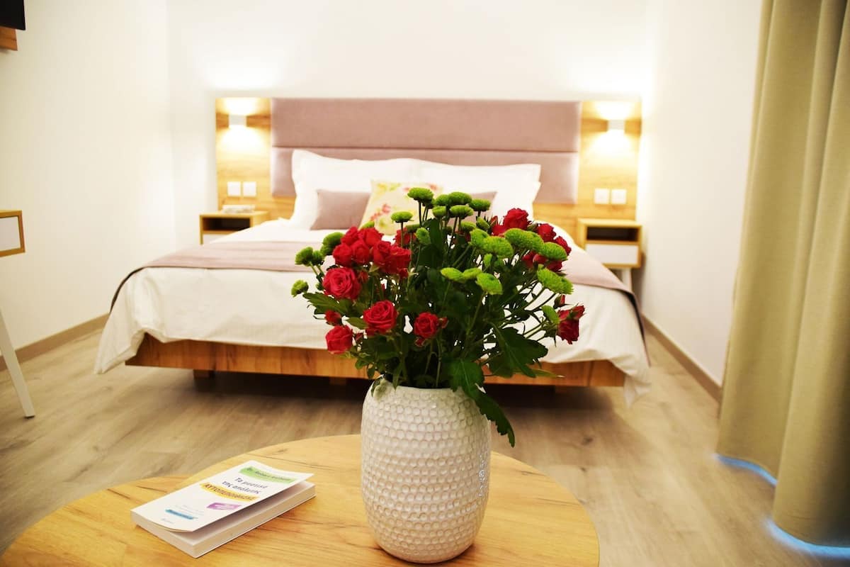 Een hotelkamer met een bed, een nachtkastje met een lamp, een vaas met rode bloemen en een brochure op het nachtkastje.