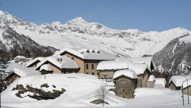 Un village de montagne enneigé, avec des bâtiments de style chalet traditionnel, un ciel bleu dégagé et, en arrière-plan, des sommets enneigés.