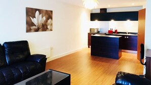 A modern living room with a black leather sofa, a glass coffee table, and a wooden floor. A kitchen with a central island and a hanging light fixture is visible in the background. - Birmingham Serviced Apartment- Ryland (Birmingham)